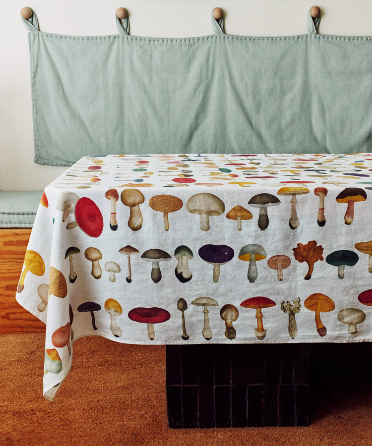 A table draped with the John Derian Fungi Tablecloth, displaying vintage mushroom illustrations, sits before a cushioned bench with a light green backrest, capturing John Derians charming style.