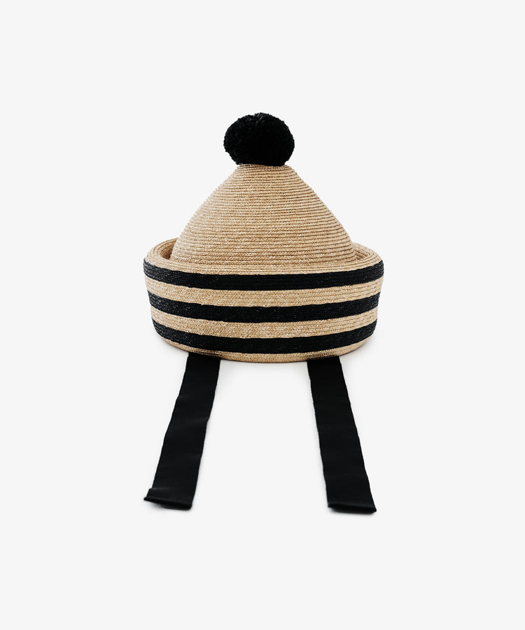 The Circus Hat is a woven, cone-shaped natural straw accessory featuring a striped crown, black pom-pom top, and two long black ties hanging down the front.