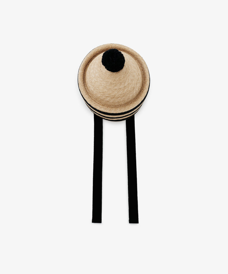The Circus Hat is a beige, round natural straw hat with a striped crown, a black pom-pom on top, and two long black ties hanging down.