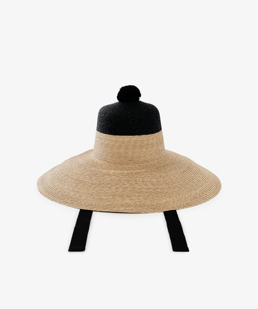 The Dahlia Hat is a handwoven straw hat with a wide brim, black crown, bold pom-pom top, and black chin strap ties, shown on a white background—a striking choice for the garden.