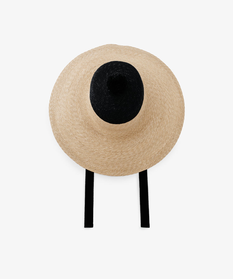 The Dahlia Hat is a handwoven straw hat with a dramatic wide brim, black crown, and elegant black ties, shown from above on a plain white background.