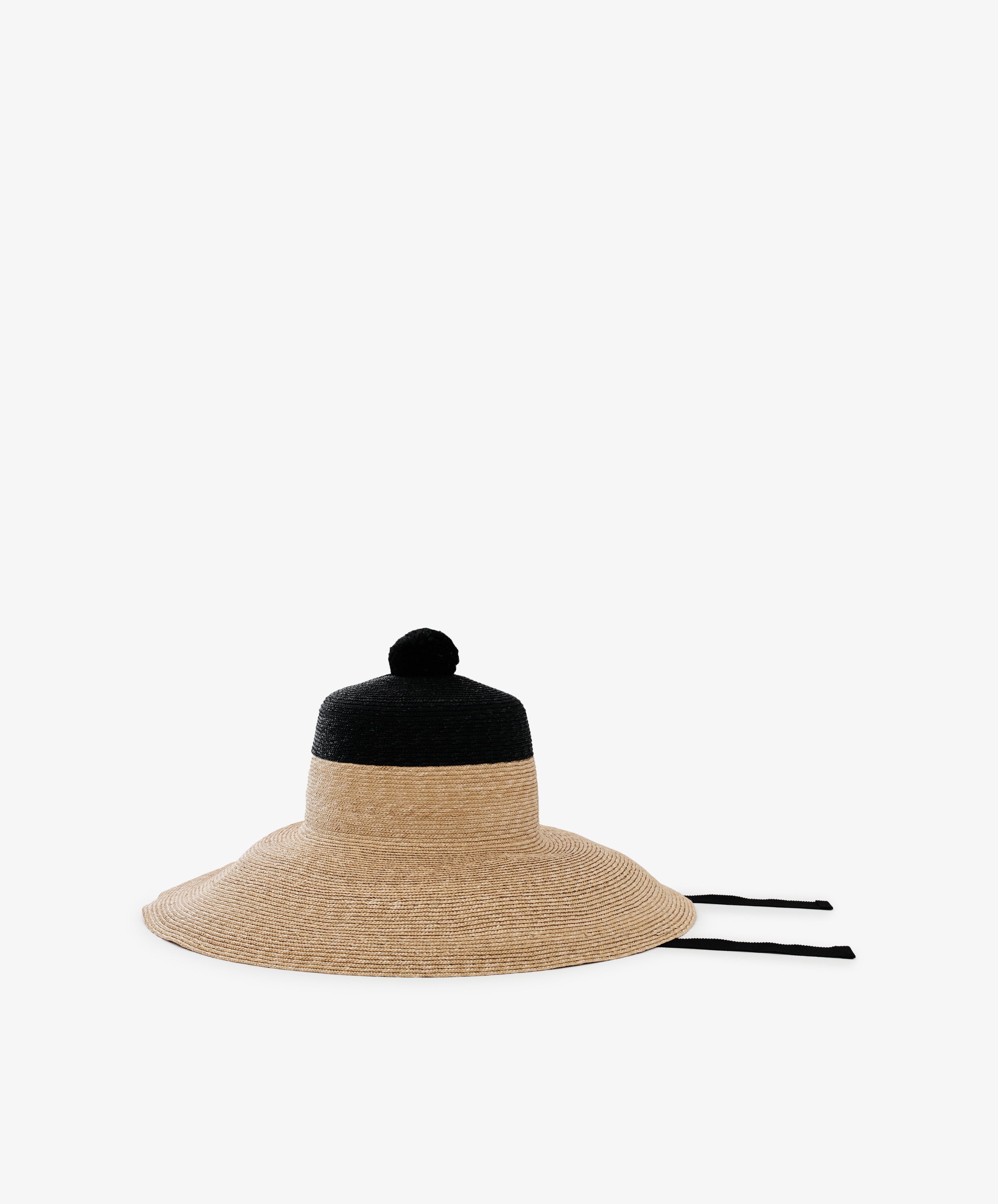 The Dahlia Hat is a handwoven straw garden hat with a natural tan lower half, black upper section, playful pom-pom, and two black ties, shown against a plain white background.