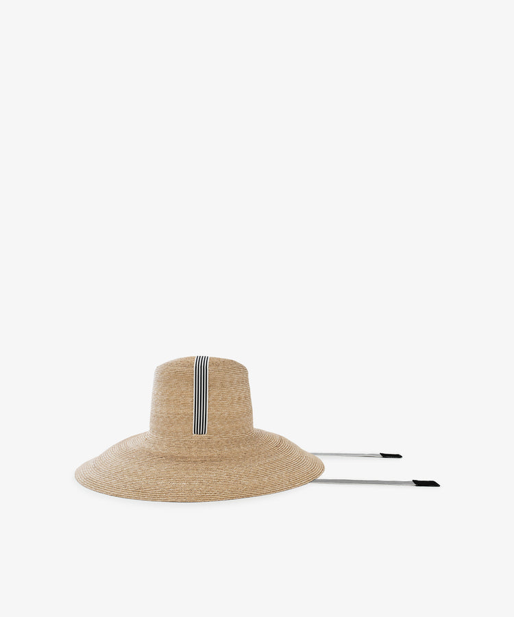 The Dahlia Hat is a natural straw hat with a wide brim, black and white striped band, and two long black-tipped chin straps, shown on a plain white background.