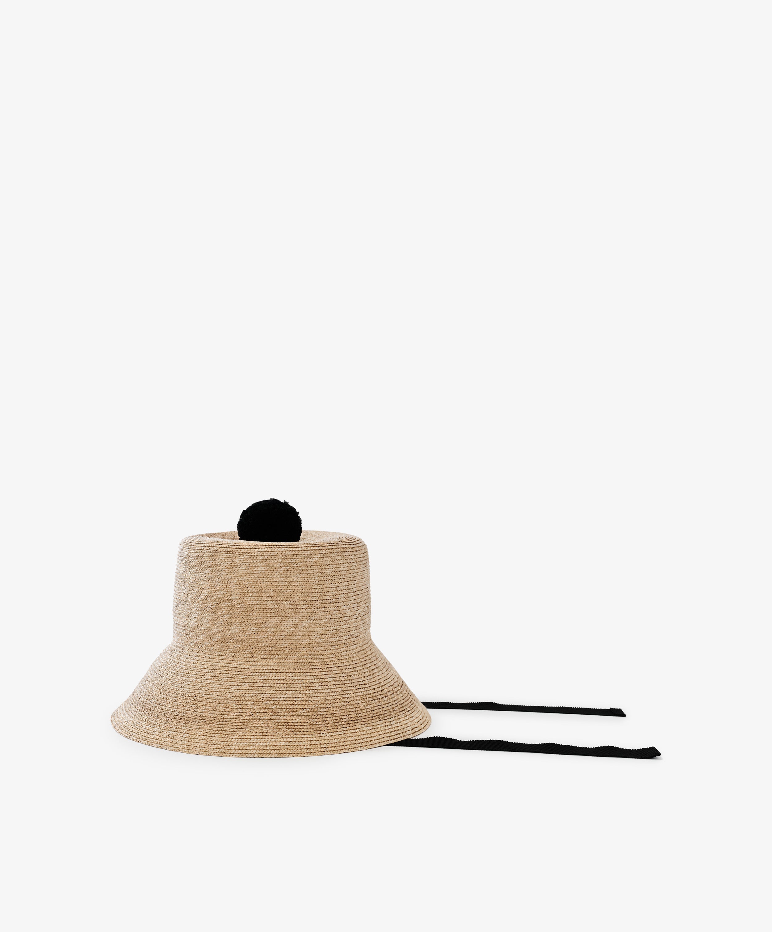 The Mount Hat, a tan handwoven straw bucket hat with a black pom-pom on top and two black tie strings, is displayed flat on a white background.