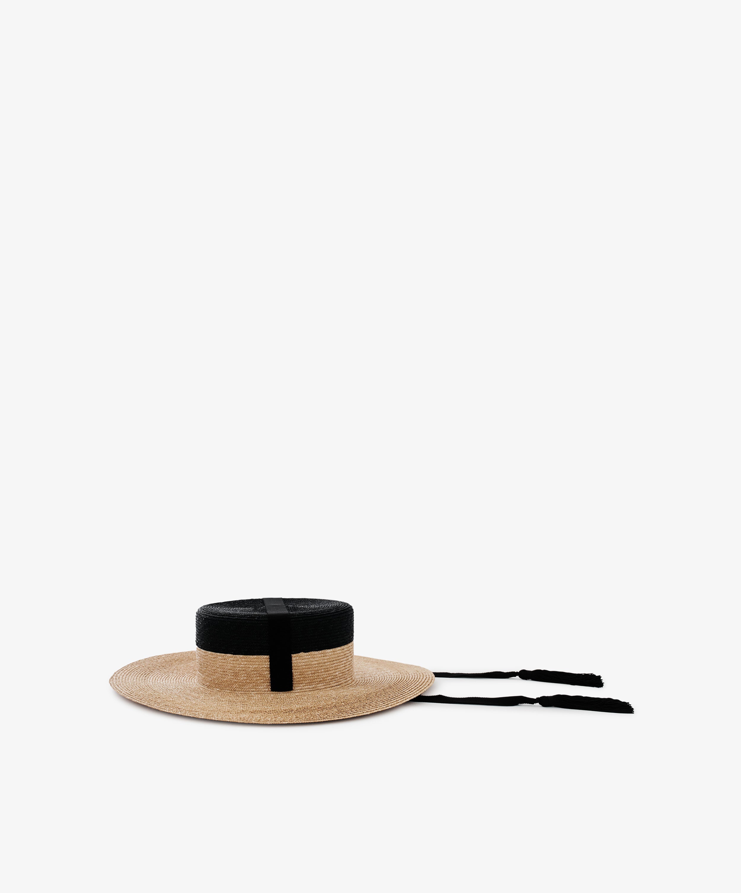 The Treaty Hat, featuring a wide brim, flat top, black band, and black ribbon ties, sits on a plain white background—offering classic resort glamour.