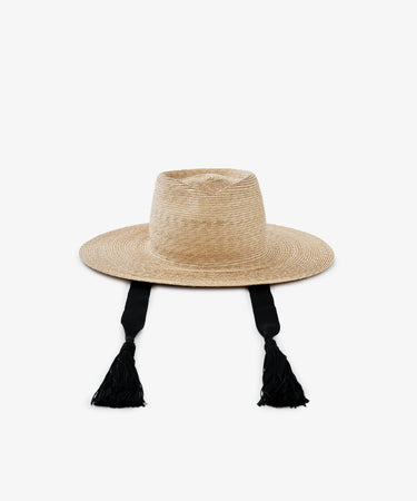 The Wosto Hat is a Mediterranean-inspired wide-brim hat with a flat crown and two long black tassel ties, providing stylish sun protection. Shown against a white background.
