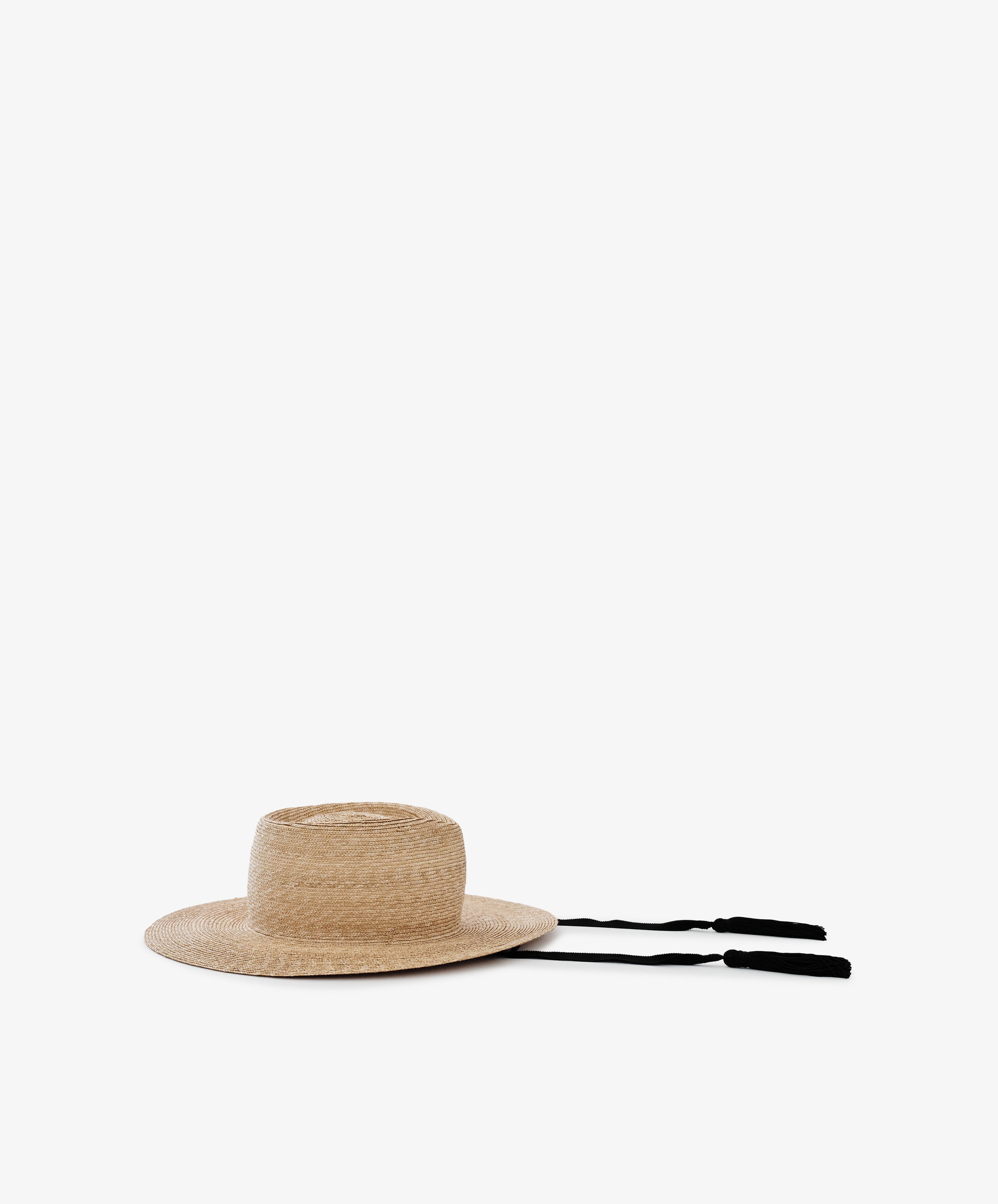 The Wosto Hat is a Mediterranean-inspired field hat with a flat crown, wide brim, and two black tassel strings, providing stylish sun protection. It is shown against a plain white background.