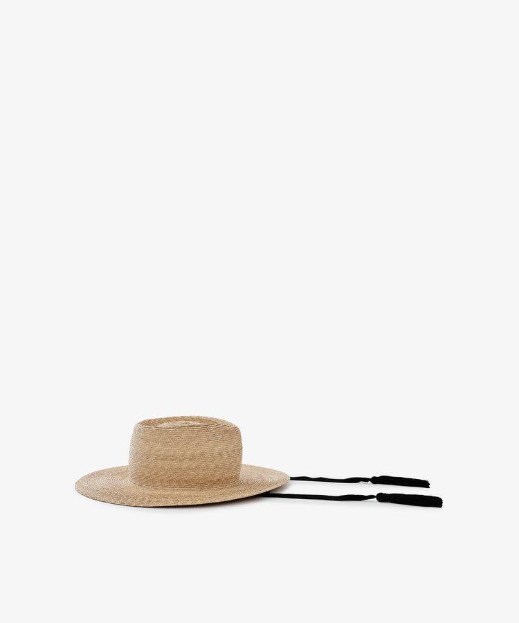 The Wosto Hat is a Mediterranean-inspired field hat with a flat crown, wide brim, and two black tassel strings, providing stylish sun protection. It is shown against a plain white background.