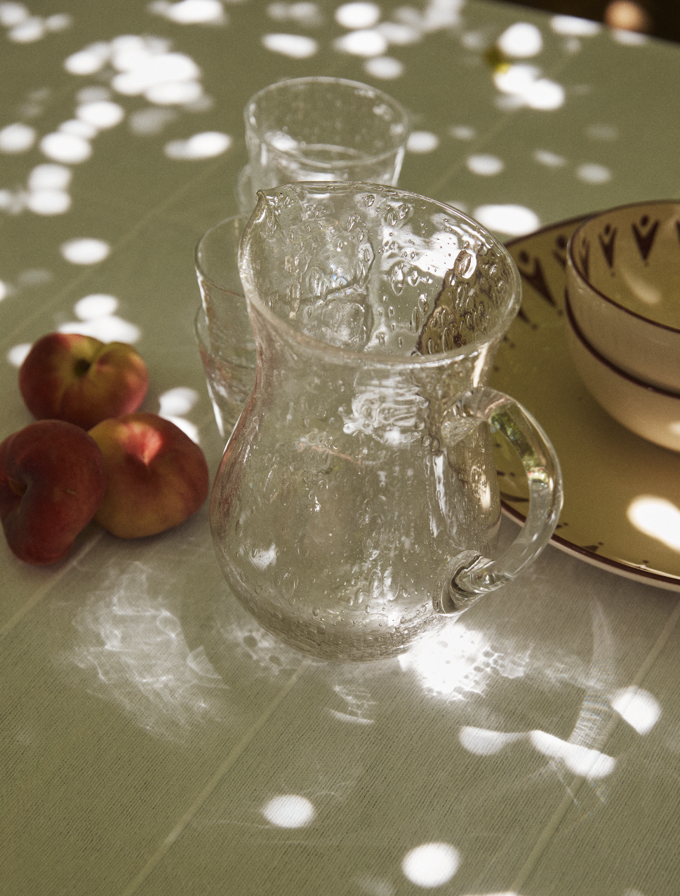 The Lauren Water Jug sits on a sunlit table beside glasses, three peaches, and patterned bowls, showcasing elegant French glassware.