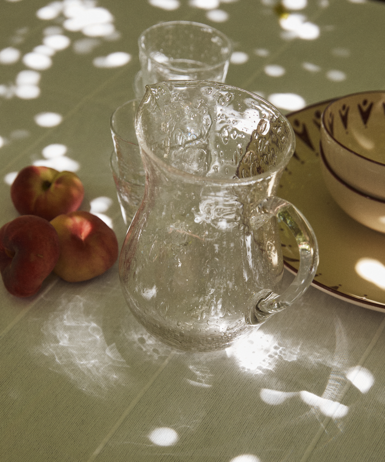 The Lauren Water Jug sits on a sunlit table beside glasses, three peaches, and patterned bowls, showcasing elegant French glassware.