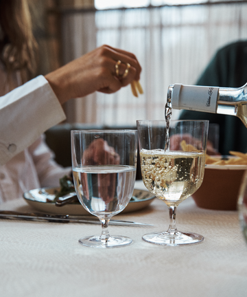 A hand reaches for food as white wine is poured into a glass beside Venezia Water Glasses, Set of 6, on a table set with refined tableware for a meal.