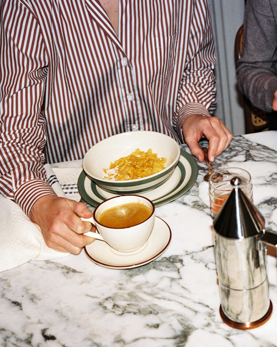 Wearing striped pajamas, a person sits at a marble table with the Guido Coffee Set—a bowl of cereal, breakfast cup of coffee, glass of juice, and metal coffee pot.