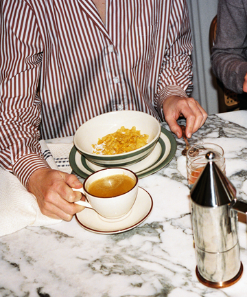 Wearing striped pajamas, a person sits at a marble table with the Guido Coffee Set—a bowl of cereal, breakfast cup of coffee, glass of juice, and metal coffee pot.