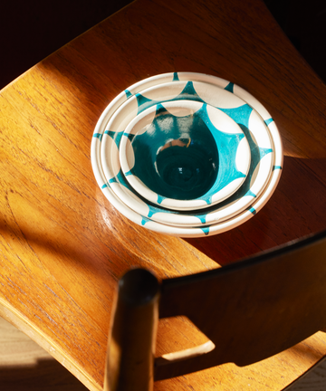 The Stella Nesting Bowls, featuring a handmade turquoise and white geometric design, rest on a wooden chair in sunlight—ideal for anyone who loves unique kitchenware.
