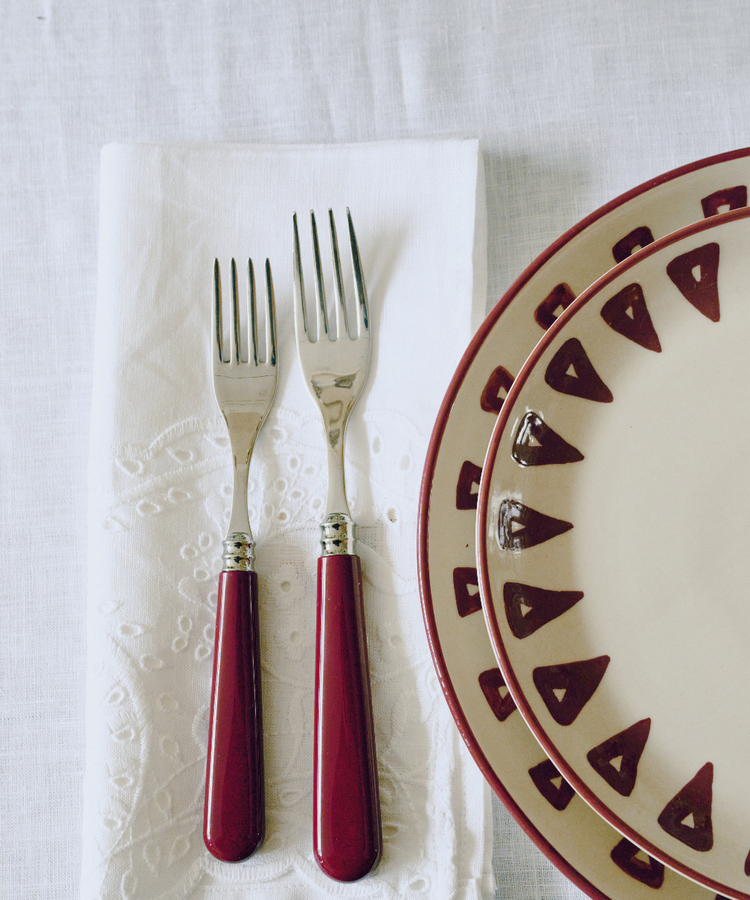 Two forks with red handles rest on a Broderie Anglaise Linen Napkin, next to stacked cream plates with red geometric patterns, all set on a crisp white tablecloth.