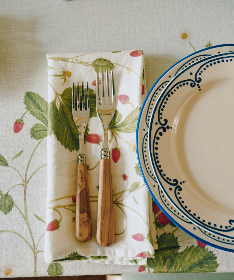 A fork and a salad fork with wooden handles rest on Fraise des Bois Linen Napkins, set of 6, decorated with wild strawberries beside a decorative plate on the table.