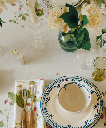 A table set with patterned plates, bowls, cutlery on a napkin, hand-blown glassware, the Elsa Candleholder, and vases with white flowers atop a floral tablecloth.