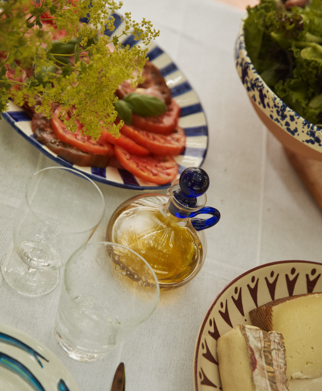 A table set with sliced tomatoes, basil, cheese, greens, two empty glasses, and the Nemo Oil Cruet with its blue stopper on a white tablecloth—a perfect display of essential kitchen accessories.