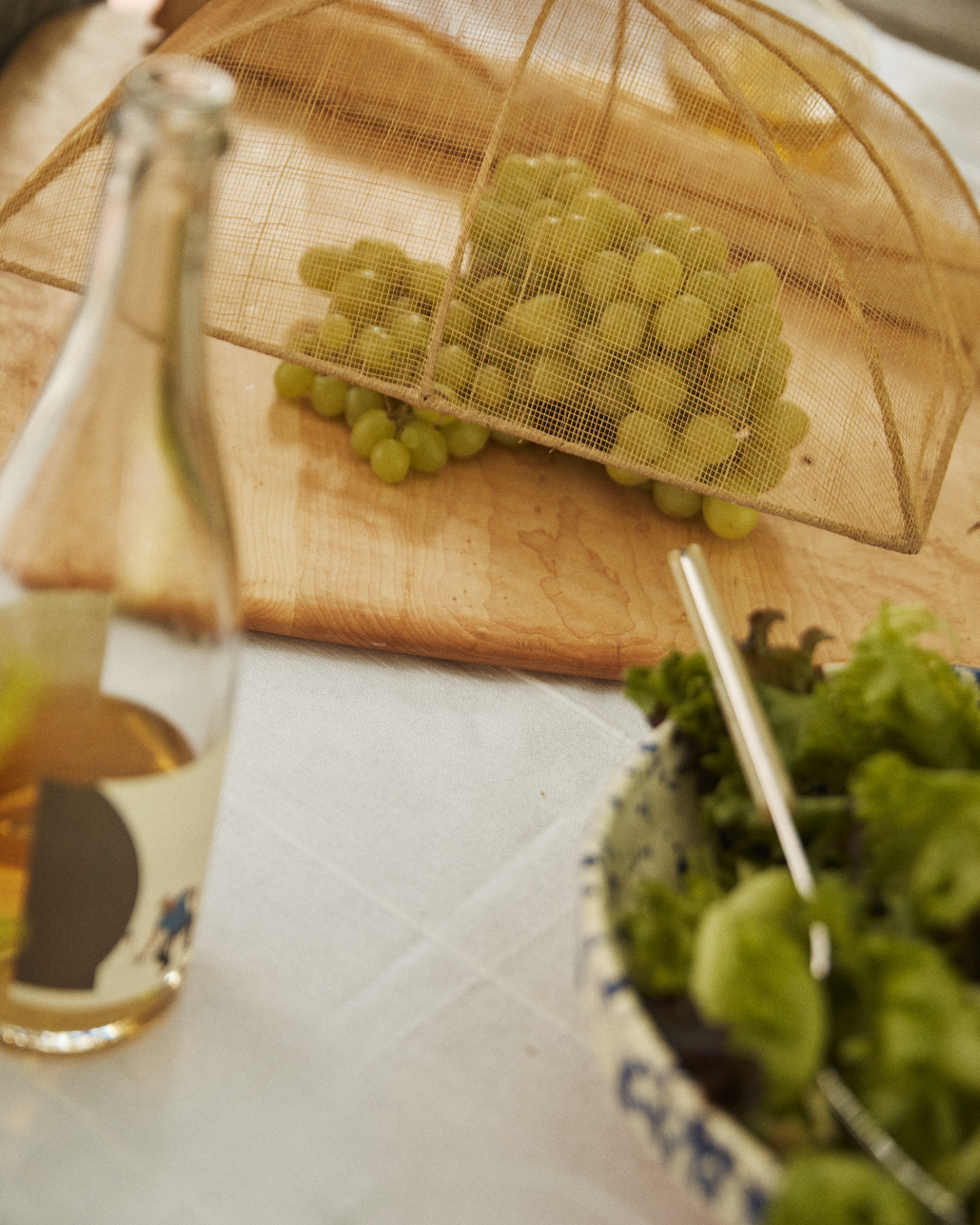A bottle, a bowl of green lettuce with a fork, and green grapes on a wooden board covered by the Jacqueline Foodcover are arranged on a table.