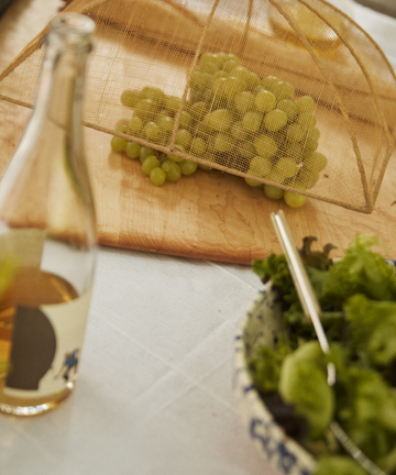 A bottle, a bowl of green lettuce with a fork, and green grapes on a wooden board covered by the Jacqueline Foodcover are arranged on a table.