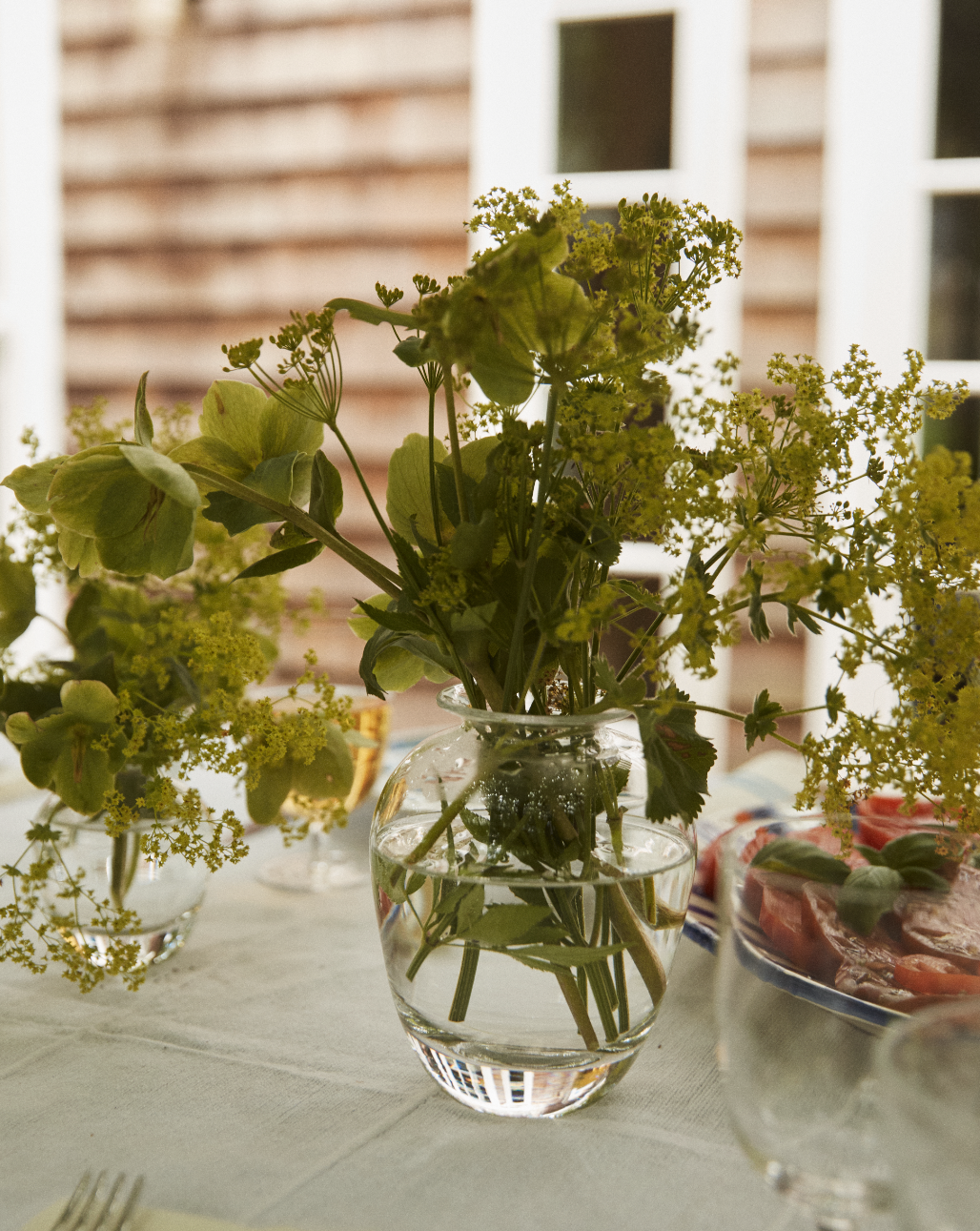 The Gem Bud Vase, hand-blown from clear glass and filled with green and yellow flowers, brings a relaxed South of France charm to a table set with dishes and glasses, against a blurred wooden wall and white-framed windows.
