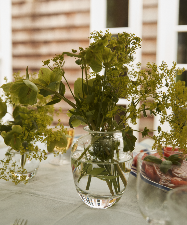 The Gem Bud Vase, hand-blown from clear glass and filled with green and yellow flowers, brings a relaxed South of France charm to a table set with dishes and glasses, against a blurred wooden wall and white-framed windows.