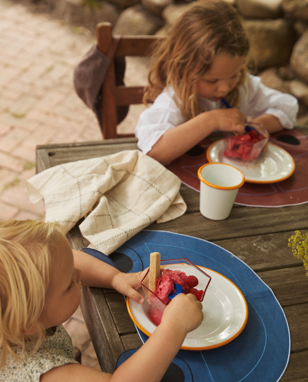 Two young children sit at a wooden outdoor table eating bowls of pink ice cream with wafer sticks. A Filial Check Napkin, Set of 6, and a cup are on the table beside them.