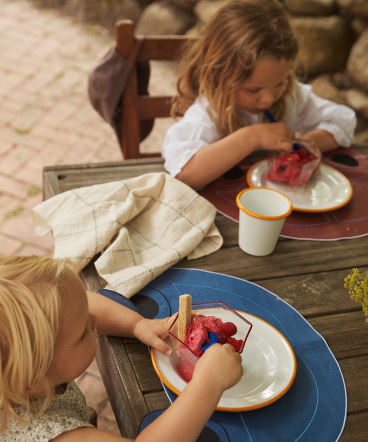 Two young children sit at a wooden outdoor table eating bowls of pink ice cream with wafer sticks. A Filial Check Napkin, Set of 6, and a cup are on the table beside them.