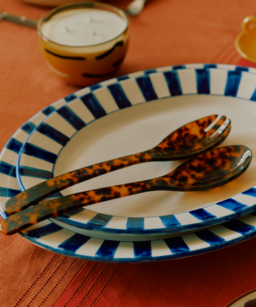 The Bestia Serving Set sits on an orange tablecloth beside two blue and white striped plates, a yellow-rimmed bowl, and a candle.
