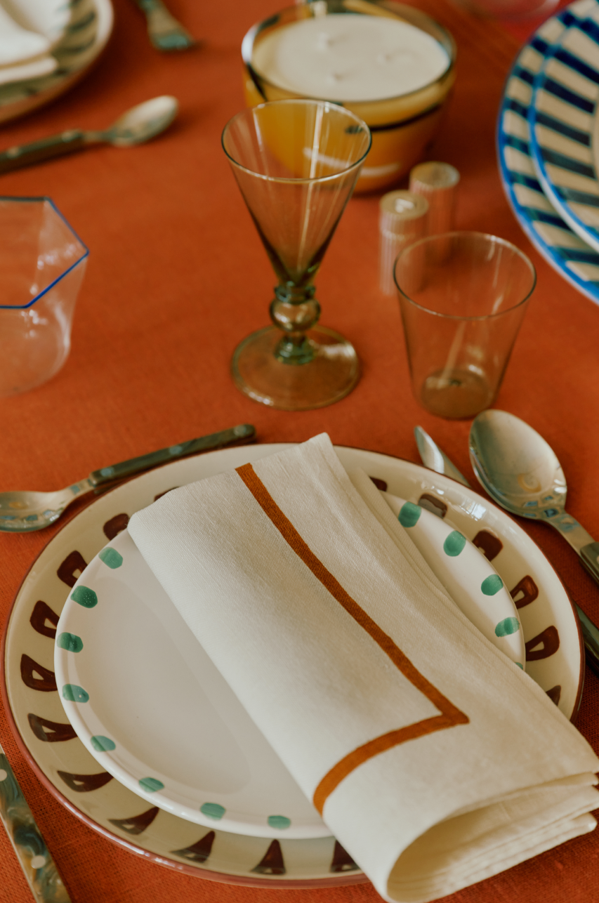 A place setting featuring a Nina Small Plate, elegant cutlery, two glasses, salt and pepper shakers, a bowl, and a folded napkin on an orange tablecloth.