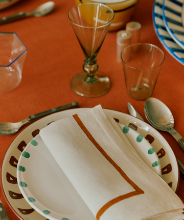 A place setting featuring a Nina Small Plate, elegant cutlery, two glasses, salt and pepper shakers, a bowl, and a folded napkin on an orange tablecloth.