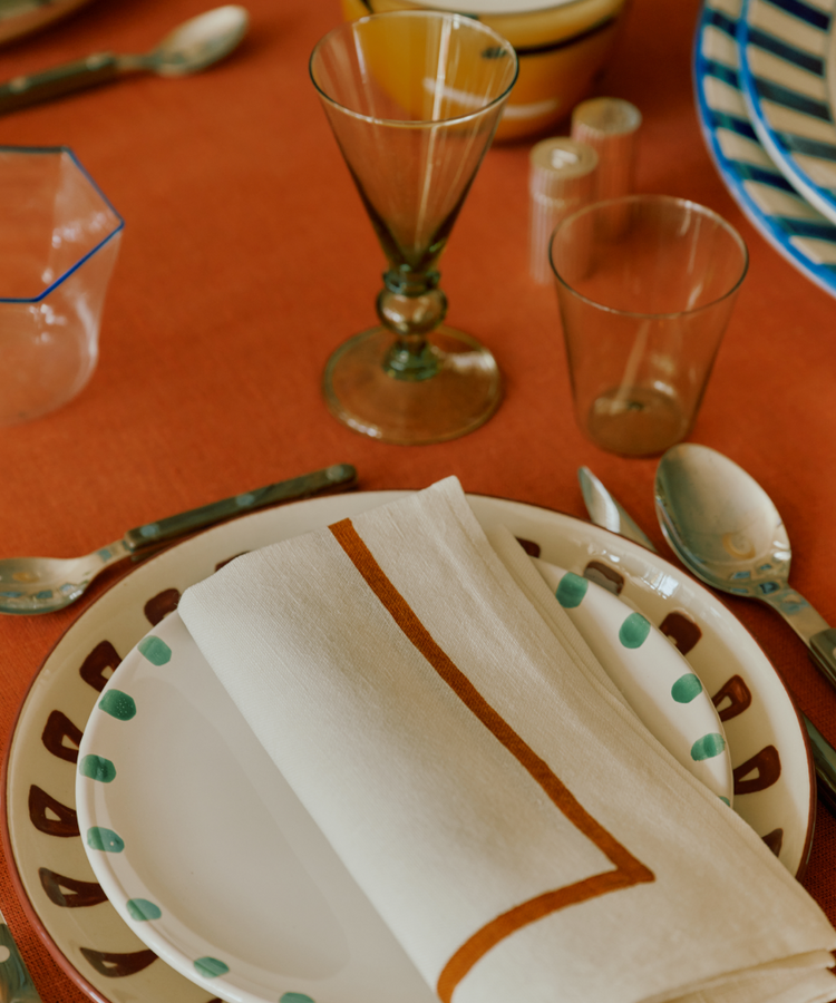 A place setting featuring a Nina Small Plate, elegant cutlery, two glasses, salt and pepper shakers, a bowl, and a folded napkin on an orange tablecloth.