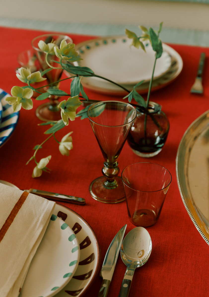 A table setting with patterned plates, silverware, elegant tableware, a napkin, a small vase with green flowers on a red tablecloth is complemented by the Beata Stemmed Wine Glass set of 6.