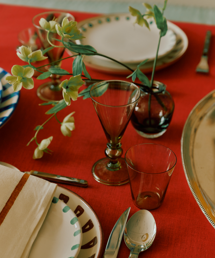 A table setting with patterned plates, silverware, elegant tableware, a napkin, a small vase with green flowers on a red tablecloth is complemented by the Beata Stemmed Wine Glass set of 6.