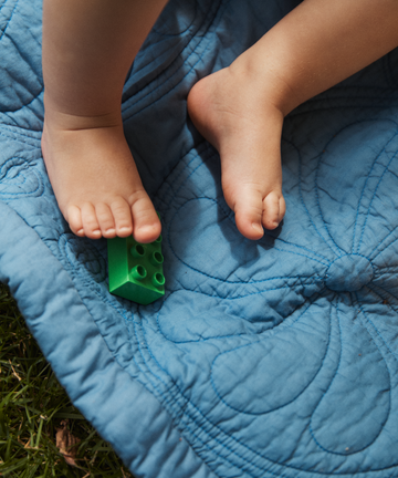 A childs bare feet rest on the Dawn Twin Bedspread, a blue double-sided quilt, with a green toy block under one foot, on grass.