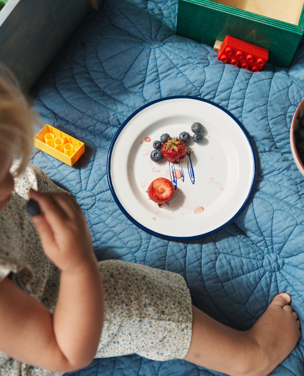 A child sits on a blue quilt next to the Il Pesce Enamel Plate, Set of 2, filled with strawberries, blueberries, and berry juice, with toy building blocks nearby.