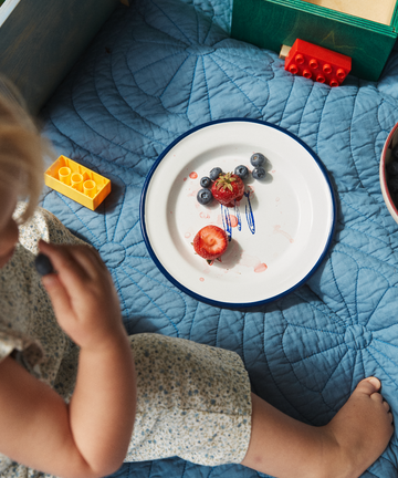A child sits on a blue quilt next to the Il Pesce Enamel Plate, Set of 2, filled with strawberries, blueberries, and berry juice, with toy building blocks nearby.