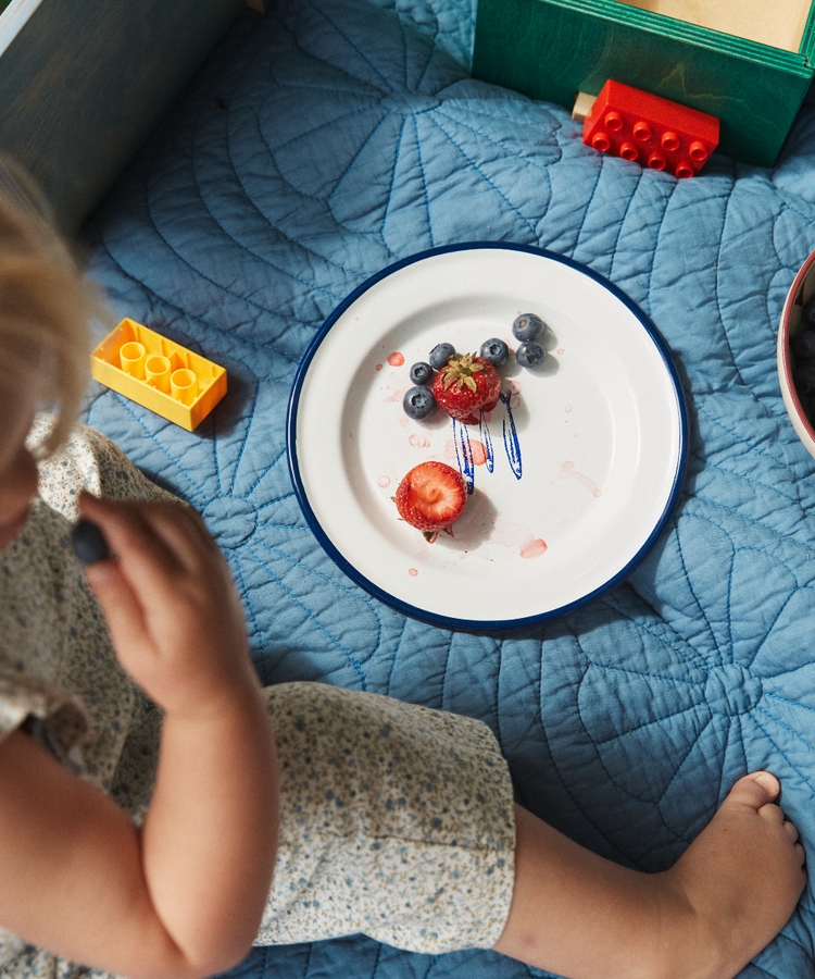 A child sits on a blue quilt next to the Il Pesce Enamel Plate, Set of 2, filled with strawberries, blueberries, and berry juice, with toy building blocks nearby.