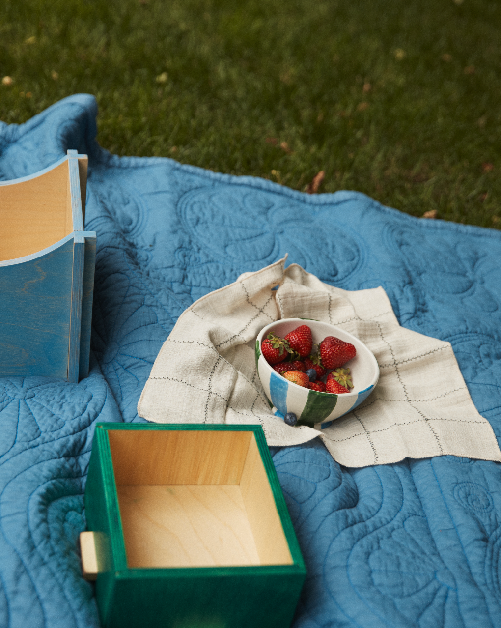 A Mina Bowl from the Set of 4 holds strawberries on a cloth, with two empty wooden boxes placed on a blue quilt spread over the grass.