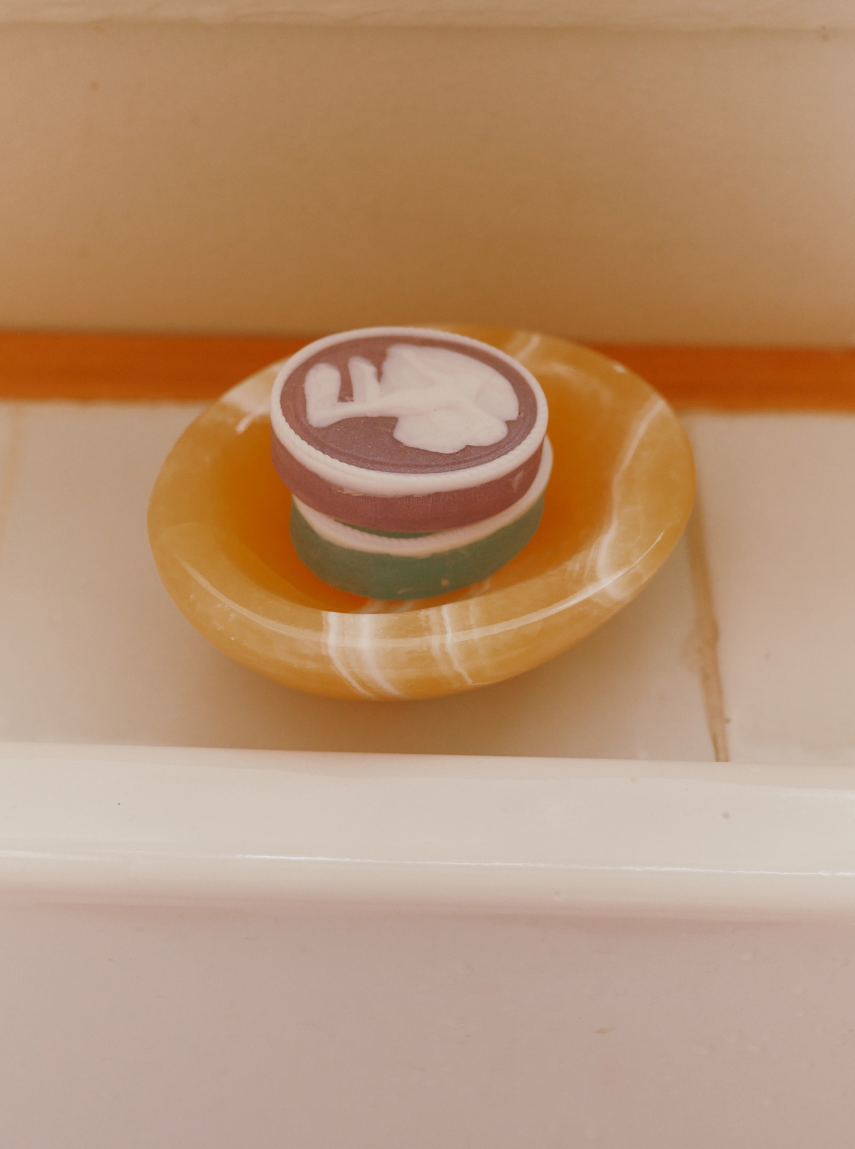 A set of two round, multicolored Diana Soaps with white designs rests in a yellow soap dish on a tiled bathroom surface, bringing elegant luxury to neoclassical decor.