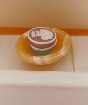 A set of two round, multicolored Diana Soaps with white designs rests in a yellow soap dish on a tiled bathroom surface, bringing elegant luxury to neoclassical decor.