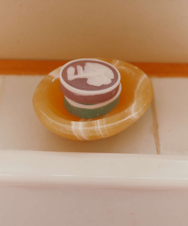 A set of two round, multicolored Diana Soaps with white designs rests in a yellow soap dish on a tiled bathroom surface, bringing elegant luxury to neoclassical decor.