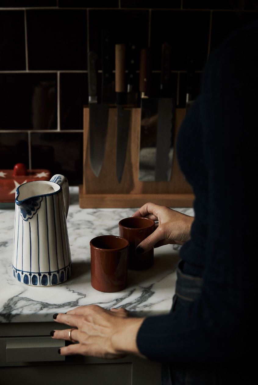 A person stands at a marble countertop with two brown cups and a green-and-white Jorge Jug with a traditional pattern; a knife block is seen in the background.