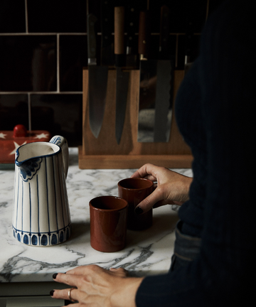 A person stands at a marble countertop with two brown cups and a green-and-white Jorge Jug with a traditional pattern; a knife block is seen in the background.