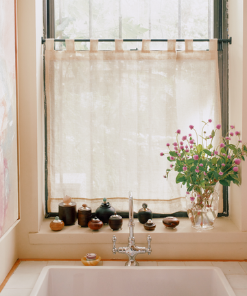 A kitchen sink beneath a window with a sheer curtain; jars, small containers, and Curiosity Box 7 line the windowsill, while a vase with pink flowers sits on the right.