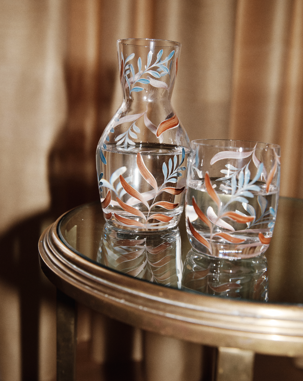 The Botanical Bedside Carafe and Glass Set, featuring a colorful leaf pattern, is displayed on a round glass table with a gold rim, both pieces filled with water.