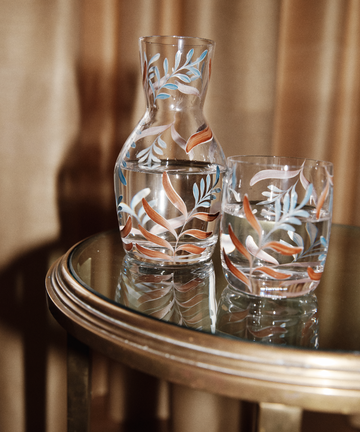 The Botanical Bedside Carafe and Glass Set, featuring a colorful leaf pattern, is displayed on a round glass table with a gold rim, both pieces filled with water.