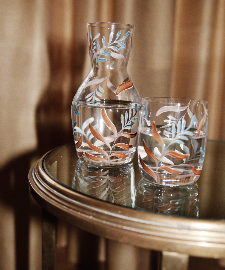 The Botanical Bedside Carafe and Glass Set, featuring a colorful leaf pattern, is displayed on a round glass table with a gold rim, both pieces filled with water.