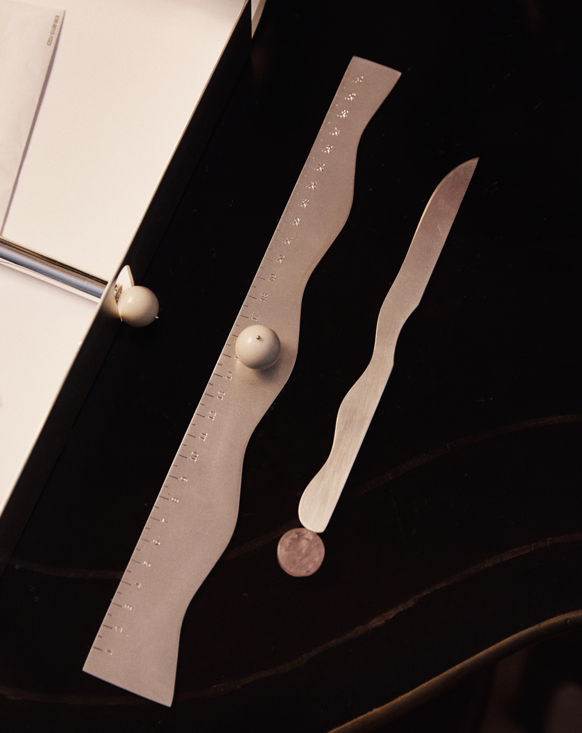 Two wavy metal Rulers, a coin, and two white spheres are arranged on a dark surface beside a white book, creating an intriguing desk accessory display.