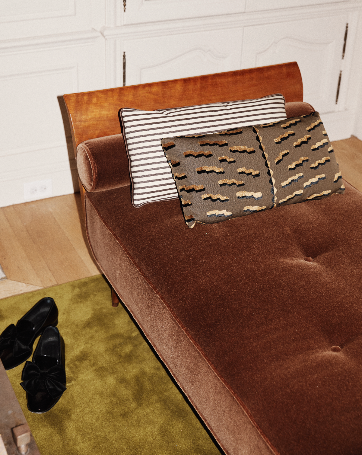 A brown upholstered chaise lounge with Polo Lumbar Cushion, black shoes on an olive green rug, and white cabinetry in the background.